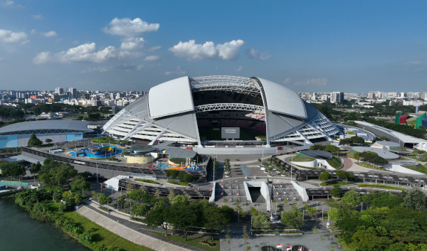 Aerial view of Singapore's National Stadium with a large, curved white roof, surrounded by green spaces and the city skyline under a blue sky.